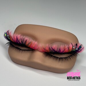May include: A pair of false eyelashes featuring a layered design with black base lashes and vibrant pink, orange, and purple wispy tips, accented with small clear rhinestones and heart-shaped charms. The text ITGIRL AESTHETICS LASH EXTENSIONS is visible.