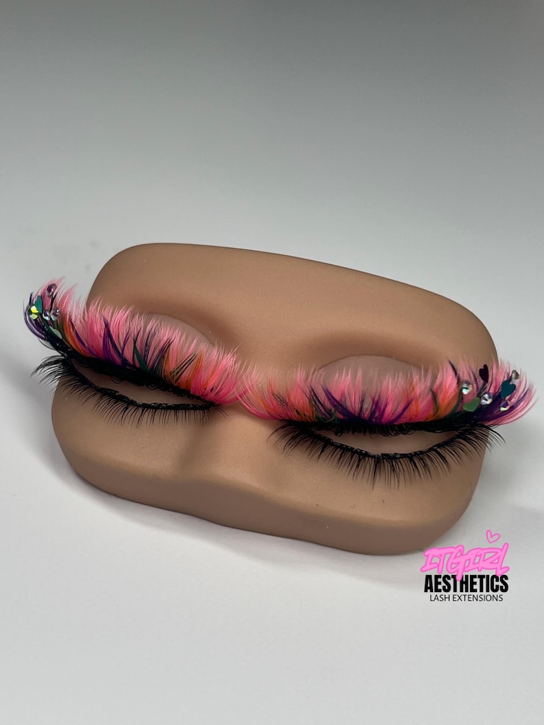 May include: A pair of false eyelashes featuring a layered design with black base lashes and vibrant pink, orange, and purple wispy tips, accented with small clear rhinestones and heart-shaped charms. The text ITGIRL AESTHETICS LASH EXTENSIONS is visible.