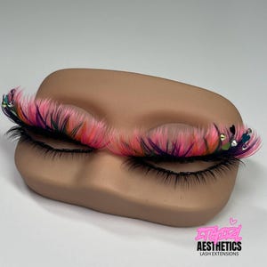 May include: False eyelashes featuring a vibrant blend of pink, purple, and orange fibers, accented with small heart-shaped charms and clear rhinestones. The lashes are displayed on a tan mannequin eye form with the text ITGIRL AESTHETICS LASH EXTENSIONS.
