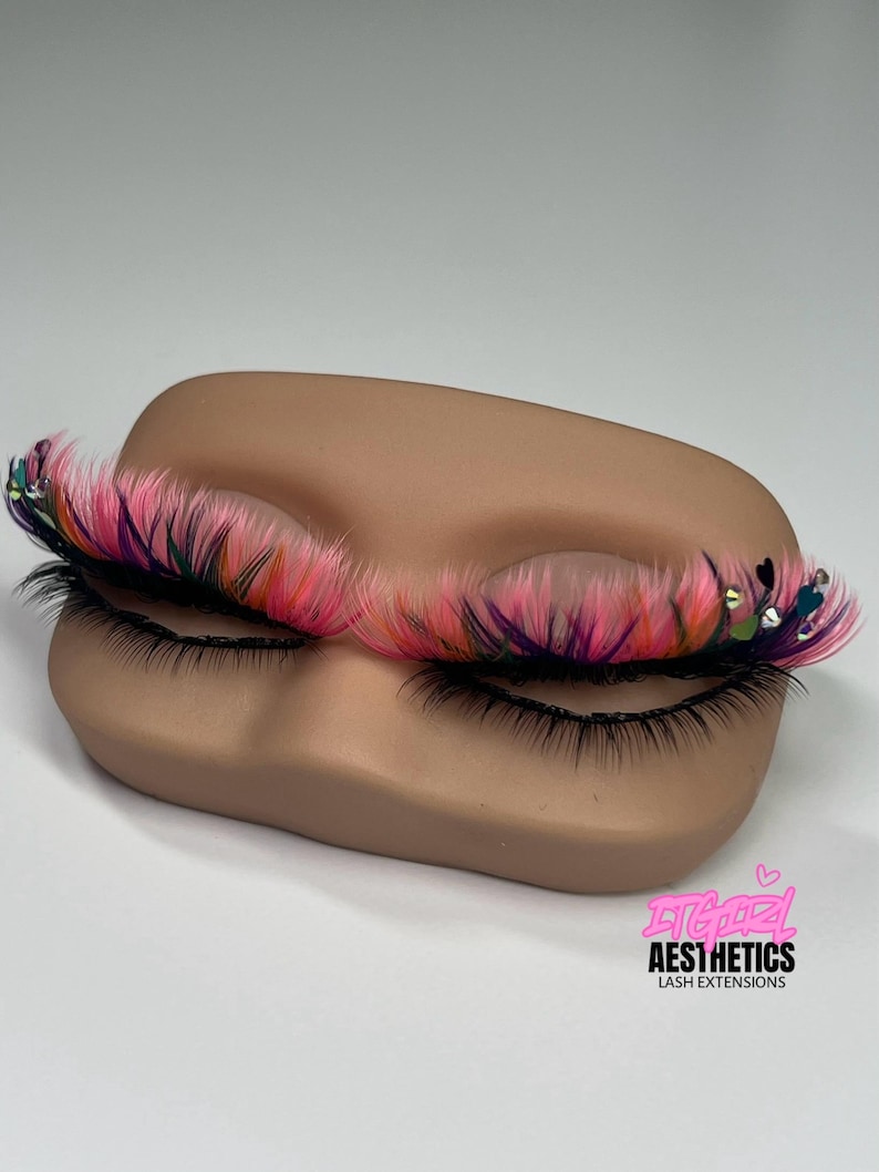 May include: False eyelashes featuring a vibrant blend of pink, purple, and orange fibers, accented with small heart-shaped charms and clear rhinestones. The lashes are displayed on a tan mannequin eye form with the text ITGIRL AESTHETICS LASH EXTENSIONS.