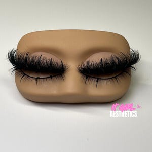 May include: A tan-coloured mannequin head with closed eyes and long, black eyelashes. The text "IT GIRL AESTHETICS" is printed in pink on the lower right corner. The background is plain white.