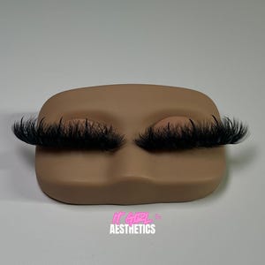 May include: A tan-coloured face-shaped display with closed eyes and long, black eyelashes. The text "IT GIRL AESTHETICS" is printed in pink and white at the bottom. The display is on a white surface.