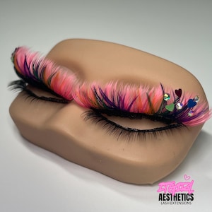 May include: A mannequin eye area featuring long, multi-colored lash extensions in shades of neon pink, purple, and black. The lashes are accented with small, iridescent heart-shaped sequins and rhinestones. The text ITGIRL AESTHETICS LASH EXTENSIONS is visible.
