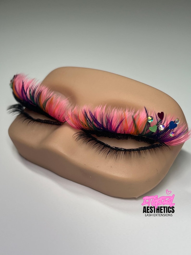 May include: A mannequin eye area featuring long, multi-colored lash extensions in shades of neon pink, purple, and black. The lashes are accented with small, iridescent heart-shaped sequins and rhinestones. The text ITGIRL AESTHETICS LASH EXTENSIONS is visible.
