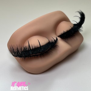 May include: A mannequin head with closed eyes and long, black eyelashes. The text "IT GIRL AESTHETICS" is printed in pink and white at the bottom left. The background is a plain, light colour.