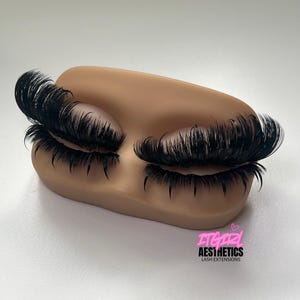 May include: A tan-coloured mannequin eye training model featuring long, dense, black synthetic lash extensions applied to both eyelids. The text ITGIRL AESTHETICS LASH EXTENSIONS with a small pink heart icon is printed in the lower right corner.