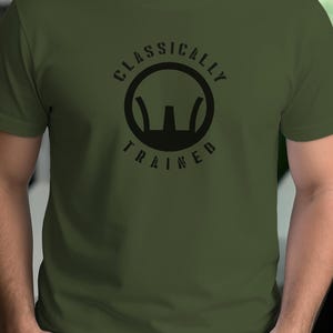 Könnte beinhalten: Olivgrünes T-Shirt mit dem schwarzen Schriftzug "CLASSICALLY TRAINED" in einem Kreis. Das kurzärmelige Shirt hat eine einfarbige Gestaltung, wobei der Text mittig auf der Brust platziert ist.