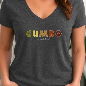 May include: Dark gray V-neck t-shirt with the word "GUMBO" in colorful letters. Below the word "GUMBO" is the word "weather" in a smaller font. The shirt is made of a soft material.