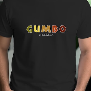 May include: Black t-shirt with the word "GUMBO" in large, colorful letters. The word "weather" is written in a smaller, cursive font below the main word. The shirt is a casual style.