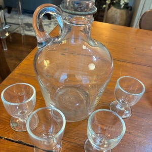 May include: A clear glass decanter with a handle and a metal stopper, alongside four small, stemmed shot glasses. The decanter has a rounded body and a curved handle. The shot glasses are small and have a rounded bowl shape.
