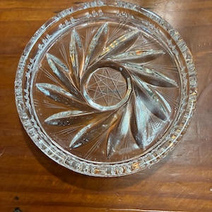 Puede incluir: Posavasos de cristal transparente con un diseño de estrella en el centro. El posavasos tiene forma circular con un patrón detallado de formas de hojas que irradian desde el centro. Los bordes son texturizados.