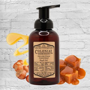 May include: A brown bottle of Colonial Merchant Deluxe-Foam Maple Nut hand soap with a black pump. The label features text and a decorative border. Caramel candies and hazelnuts are arranged around the bottle.