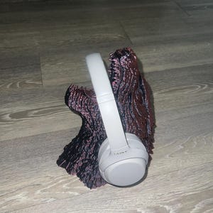 以下が含まれることがあります： 白のSony製ヘッドホンが、質感のある濃い茶色と黒の装飾スタンドに置かれています。ヘッドホンはスタンドに配置され、コントラストのある背景に対して製品のデザインと色を際立たせています。