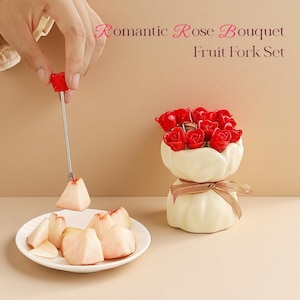Puede incluir: Un juego de tenedores para fruta con una punta en forma de rosa roja, utilizado para recoger un trozo de fruta. El juego incluye un soporte de cerámica blanca con decoraciones de rosas rojas y una cinta decorativa. El texto "Romantic Rose Bouquet Fruit Fork Set" es visible.