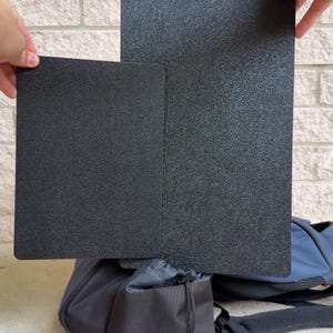 Könnte beinhalten: Zwei rechteckige schwarze Platten mit einer texturierten, körnigen Oberfläche. Die Platten werden aufrecht gehalten, wobei eine deutlich höher ist als die andere, positioniert über einem dunkelblauen und schwarzen Kordelzugbeutel mit verstellbaren Riemen.