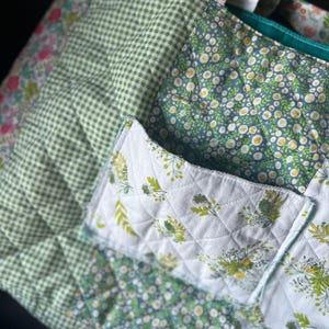 May include: A quilted tote bag with a variety of floral and gingham patterns in shades of green, white, and pink. The bag features a front pocket with a leaf design and a teal interior.