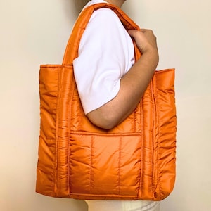 Könnte beinhalten: Eine große, gesteppte orangefarbene Tragetasche mit rechteckiger Form. Die Tasche hat zwei Henkel und eine Tasche auf der Vorderseite. Das Material hat ein glänzendes, leicht reflektierendes Aussehen. Die Tasche wird von einer Person gehalten.