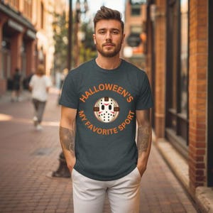 May include: Dark gray t-shirt with the text "Halloween's My Favorite Sport" in orange. The graphic features a hockey mask design. The person in the image is wearing white pants and standing on a brick-paved street.