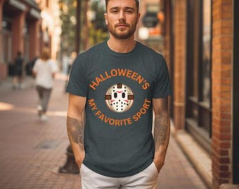May include: Dark gray t-shirt with the text "Halloween's My Favorite Sport" in orange. The graphic features a hockey mask design. The person in the image is wearing white pants and standing on a brick-paved street.