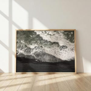 Op de afbeelding: Een rechthoekige wandkunstprint met een luchtfoto van een donker zwart zandstrand dat het witte schuim van beukende oceaangolven ontmoet. De afbeelding is ingelijst in een lichtgekleurde houten lijst en toont een contrastrijk monochroom zeegezicht.