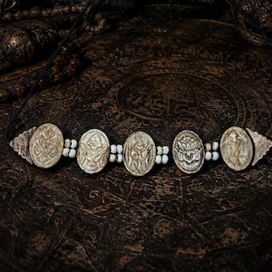 May include: A necklace featuring five oval-shaped, off-white carved pendants depicting symbols including a lotus and knots, separated by small white beads. The pendants are strung on a thin black cord with black tassels at the ends.