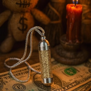 Puede incluir: Un collar plateado con un colgante cilíndrico lleno de purpurina dorada descansa sobre dos billetes de dólar. Una muñeca de arpillera con ojos de botón y un signo de dólar está en el fondo, junto con una vela encendida.