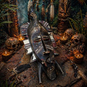 May include: A carved wooden mask with a bird-like crest and geometric patterns in red, white, and black. The mask rests on a textured rock surface, surrounded by candles, skulls, and feathers, creating a mystical, tribal aesthetic.
