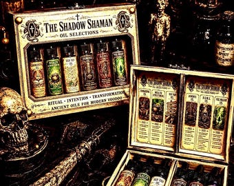 Puede incluir: Un conjunto de selecciones de aceites The Shadow Shaman en dos cajas de madera. Cada caja contiene seis frascos de vidrio pequeños etiquetados como Commanding, Wealth Attraction, Van Van, Dominus, D.U.M.E. y Veil Walker, con ilustraciones y texto detallados.