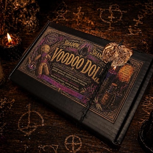 Puede incluir: Una caja negra con letras doradas y una ilustración de muñeca vudú. La caja está etiquetada como "Shadow Shaman Voodoo Doll" e incluye un sello de cera y una botella de "Voodoo Oil". Velas encendidas y una calavera en el fondo.