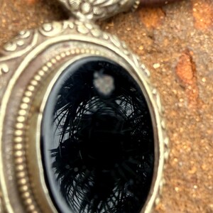 May include: Close-up of a silver-toned pendant with an oval, black gemstone. The pendant has an ornate, detailed design around the gemstone and a brown leather cord. The gemstone reflects a palm tree.