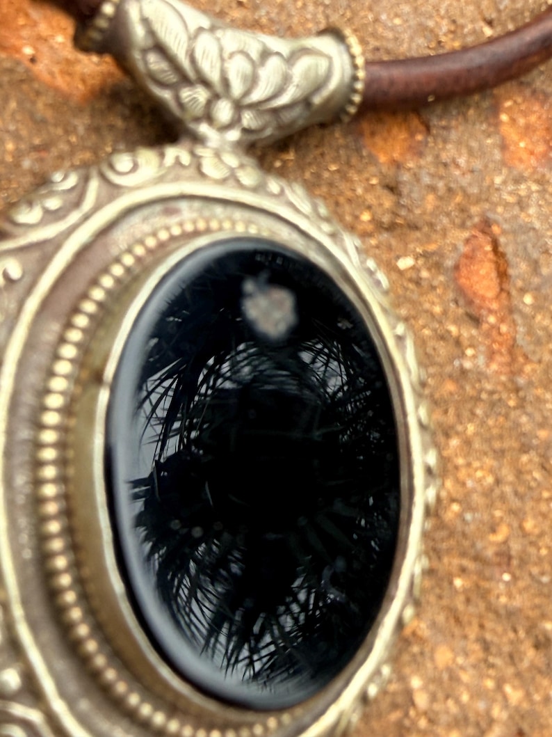 May include: Close-up of a silver-toned pendant with an oval, black gemstone. The pendant has an ornate, detailed design around the gemstone and a brown leather cord. The gemstone reflects a palm tree.