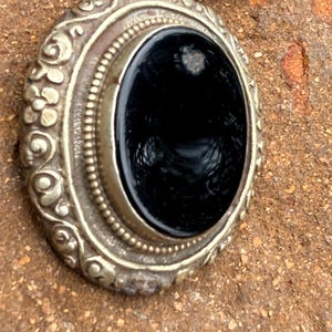 May include: Close-up of an ornate silver pendant with a large, oval black stone. The pendant has detailed floral and beaded designs around the stone. A brown leather cord is attached to the pendant.