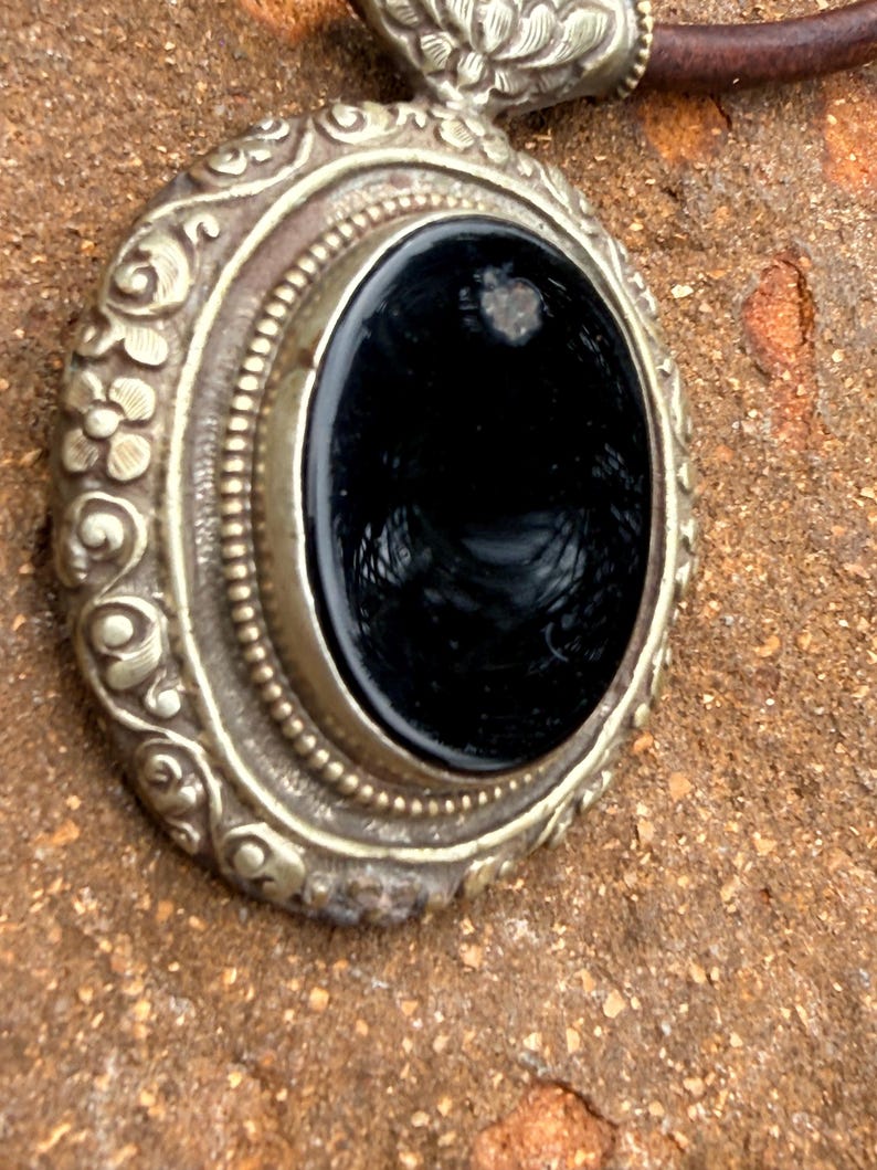May include: Close-up of an ornate silver pendant with a large, oval black stone. The pendant has detailed floral and beaded designs around the stone. A brown leather cord is attached to the pendant.