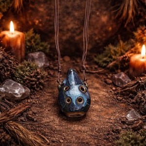 May include: A blue and green ceramic ocarina pendant with multiple holes and a brown leather cord. The pendant is set against a natural, earthy background with candles and foliage, creating a rustic aesthetic.