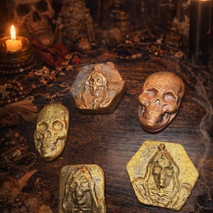 Puede incluir: Una variedad de objetos decorativos con temática de calaveras y la muerte, en oro y bronce. Los objetos incluyen objetos con forma de calavera y piezas hexagonales y cuadradas con diseños de la muerte. Velas encendidas y otros objetos ocultos en el fondo.