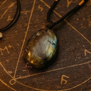 May include: A labradorite pendant necklace with a black cord. The oval-shaped stone has a dark, iridescent surface with flashes of blue and gold. The necklace rests on a dark wooden surface with etched symbols.