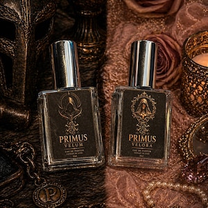 Può includere: Due flaconi di profumo rettangolari con tappi argentati ed etichette scure, etichettati "PRIMUS VELUM" e "PRIMUS VELORA". I flaconi sono appoggiati su una superficie strutturata con una maschera, rose e gioielli sullo sfondo. I flaconi sono pieni di profumo.