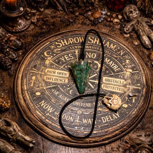 May include: A divination set with a wooden Ouija board, a green crystal pendulum, and a skull pendant. The board has a circular design with the words "Shadow Shaman" and "Yes, No, Maybe". The scene is set on a dark wooden surface.