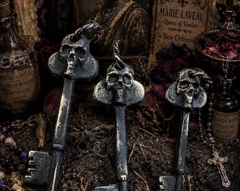 Puede incluir: Tres llaves maestras de metal oscuro y desgastado de diferentes tamaños, cada una con un motivo de calavera detallado en la cabeza. Las llaves tienen un acabado envejecido con toques dorados y plateados, e incluyen un aro de metal trenzado.