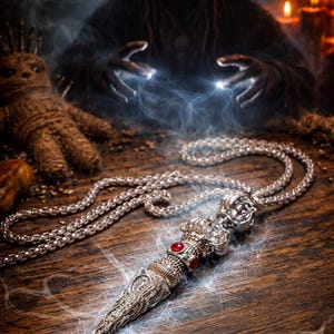 Op de afbeelding: Een zilveren hangerketting met een dolkvormige bedel, versierd met rode edelstenen. De ketting ligt op een houten oppervlak, met een mystieke achtergrond met een figuur en kaarsen. De hanger is ingewikkeld gedetailleerd.