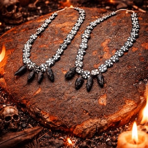 May include: Two necklaces with black and white floral-patterned beads and black pendant charms. The necklaces are displayed on a textured, brown surface, with candles, skulls, and flames, evoking a gothic or occult theme.
