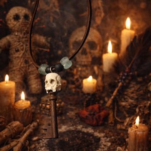May include: A pendant necklace with a skull charm and a vintage key. The necklace hangs from a brown cord, with blue and black beads. The background includes candles, a voodoo doll, and skulls.