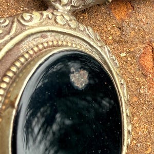 May include: Close-up of a silver-toned pendant with an oval, black stone center. The pendant has ornate detailing around the stone and a brown leather cord. The background is a textured, brown surface.