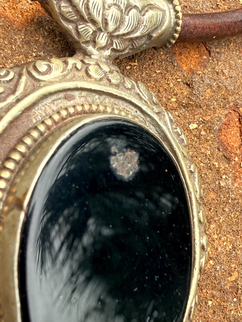 May include: Close-up of a silver-toned pendant with an oval, black stone center. The pendant has ornate detailing around the stone and a brown leather cord. The background is a textured, brown surface.
