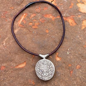 May include: A silver pendant necklace with a round, ornate pendant featuring floral and scroll designs. The pendant is attached to a dark brown leather cord necklace. The necklace is displayed on a textured, reddish-brown surface.