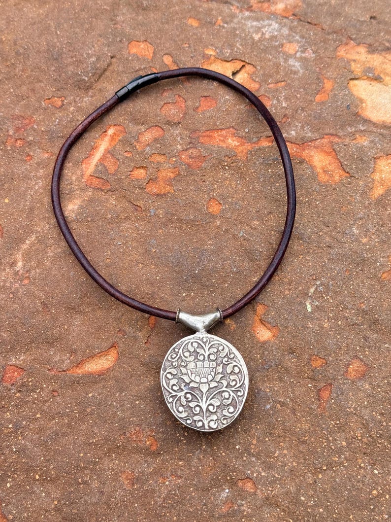 May include: A silver pendant necklace with a round, ornate pendant featuring floral and scroll designs. The pendant is attached to a dark brown leather cord necklace. The necklace is displayed on a textured, reddish-brown surface.