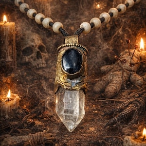 May include: A pendant necklace featuring a clear crystal point and a black stone set in an ornate gold-coloured metal casing. The necklace has a beaded strand. Lit candles and a skull are in the background, creating a mystical theme.