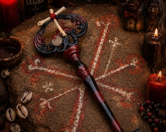 Puede incluir: Una llave decorativa con acabado degradado en negro y rojo, un mango ornamentado con una pequeña cruz de hueso y una moneda. La llave descansa sobre una piedra marcada con un símbolo de cruz blanca y roja, rodeada de velas y figuras.