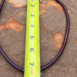 May include: A brown leather necklace with a silver pendant. The necklace is measured with a yellow tape measure, showing a length of approximately 8 inches. The pendant has an ornate design.