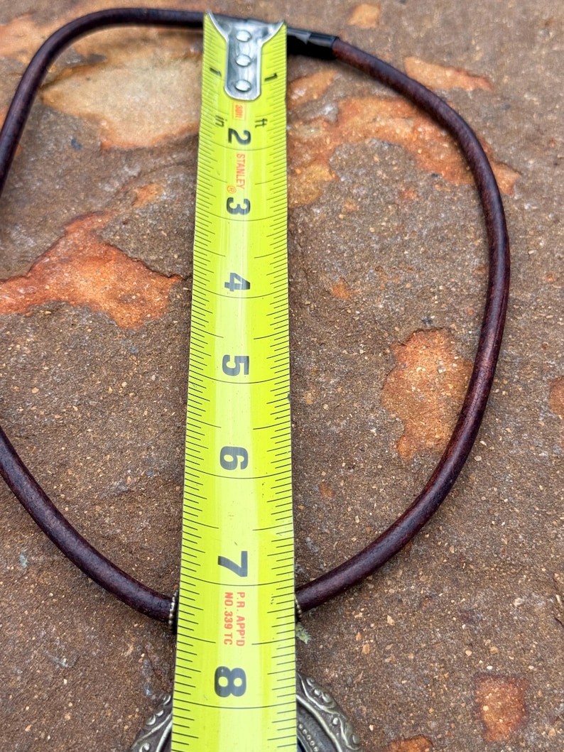 May include: A brown leather necklace with a silver pendant. The necklace is measured with a yellow tape measure, showing a length of approximately 8 inches. The pendant has an ornate design.